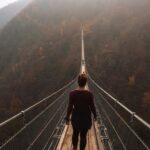 A woman with her back to the camera stands on a high, narrow suspension bridge