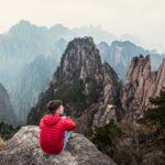 A man in a red jacket sits on the edge of a cliff looking towards mountains