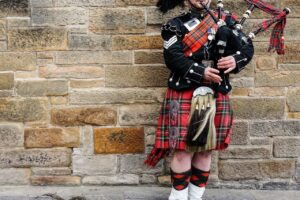 Scottish bagpiper in full Highland dress playing the pipes against a stone wall during a Highland Games event.
